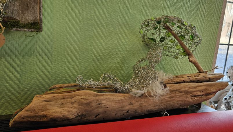 Eine Skulptur aus Treibholz vom Rheinufer, auf dem aus Perlen und Draht geformt eine Mensch unter einem Schirm liegt.