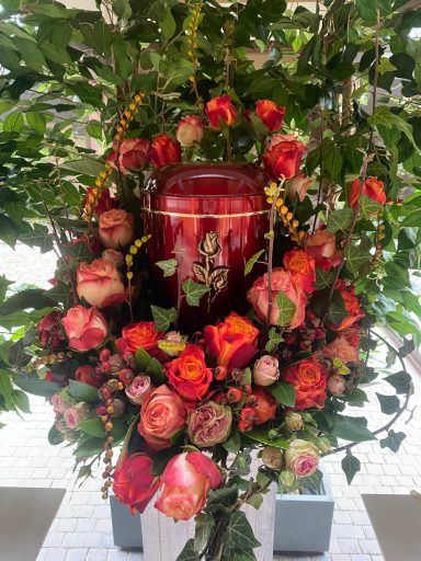 Urnengesteck mit facettenreichen roten Rosen und grünem aufgestelltem Blätterkragen um die Urne herum gesteckt.