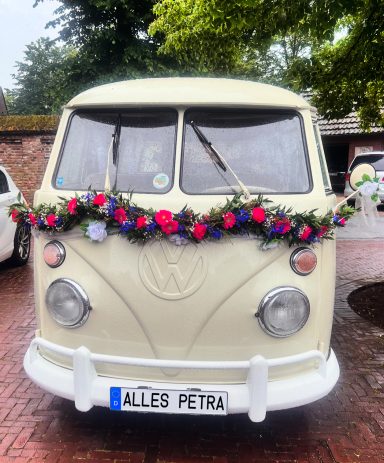 VW Bulli in elfenbeiweiß mit rot grüner Blumengirlande verziehrt für Hochzeit.